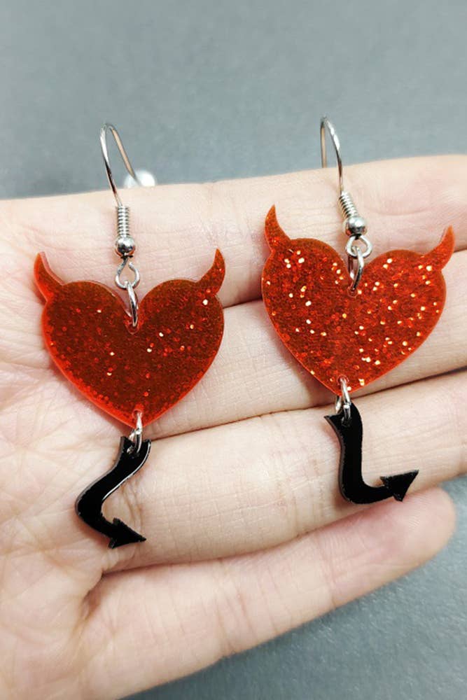 Heart-shaped earrings with devil horns and tails held in a hand against a gray background