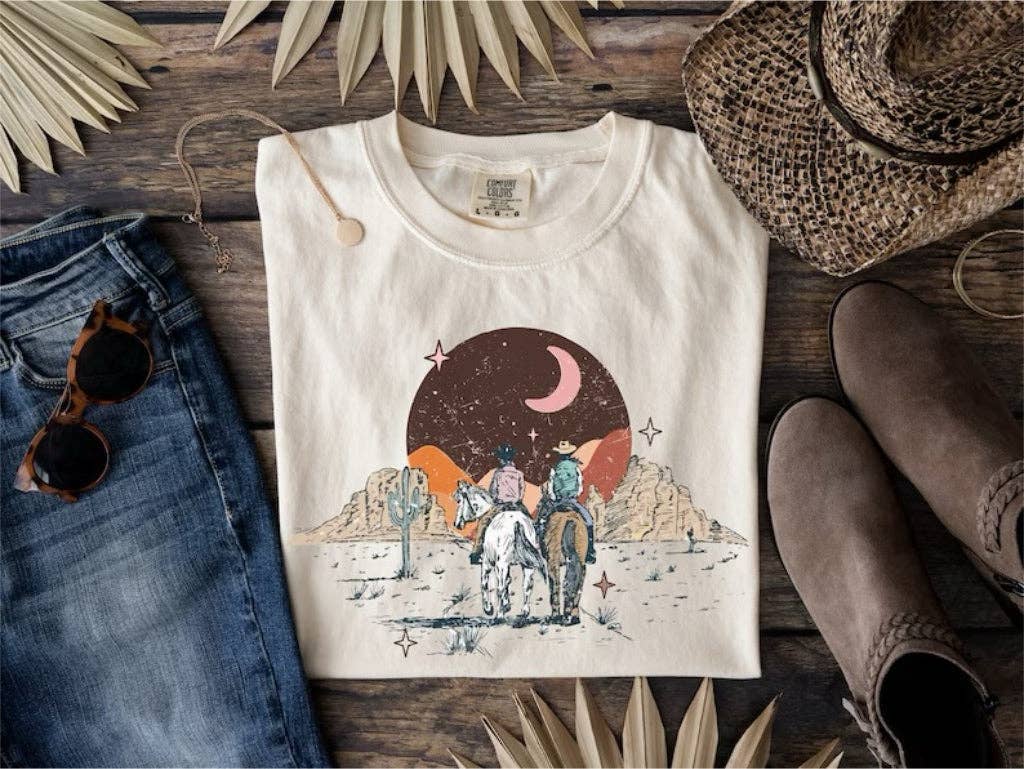 White t-shirt with desert scene graphic on a wooden surface with jeans, boots, and hat.