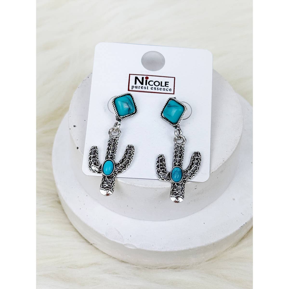 Cactus-shaped earrings with turquoise stones on a white card with 'Nicole' branding, displayed on a white surface.