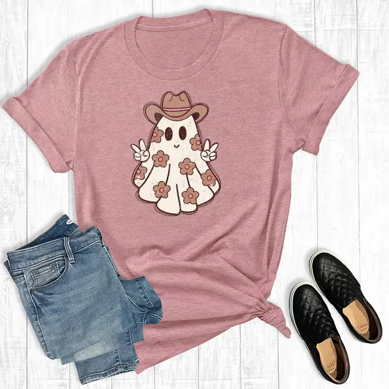 Pink t-shirt with a ghost wearing a cowboy hat design, paired with jeans and shoes on a wooden surface.