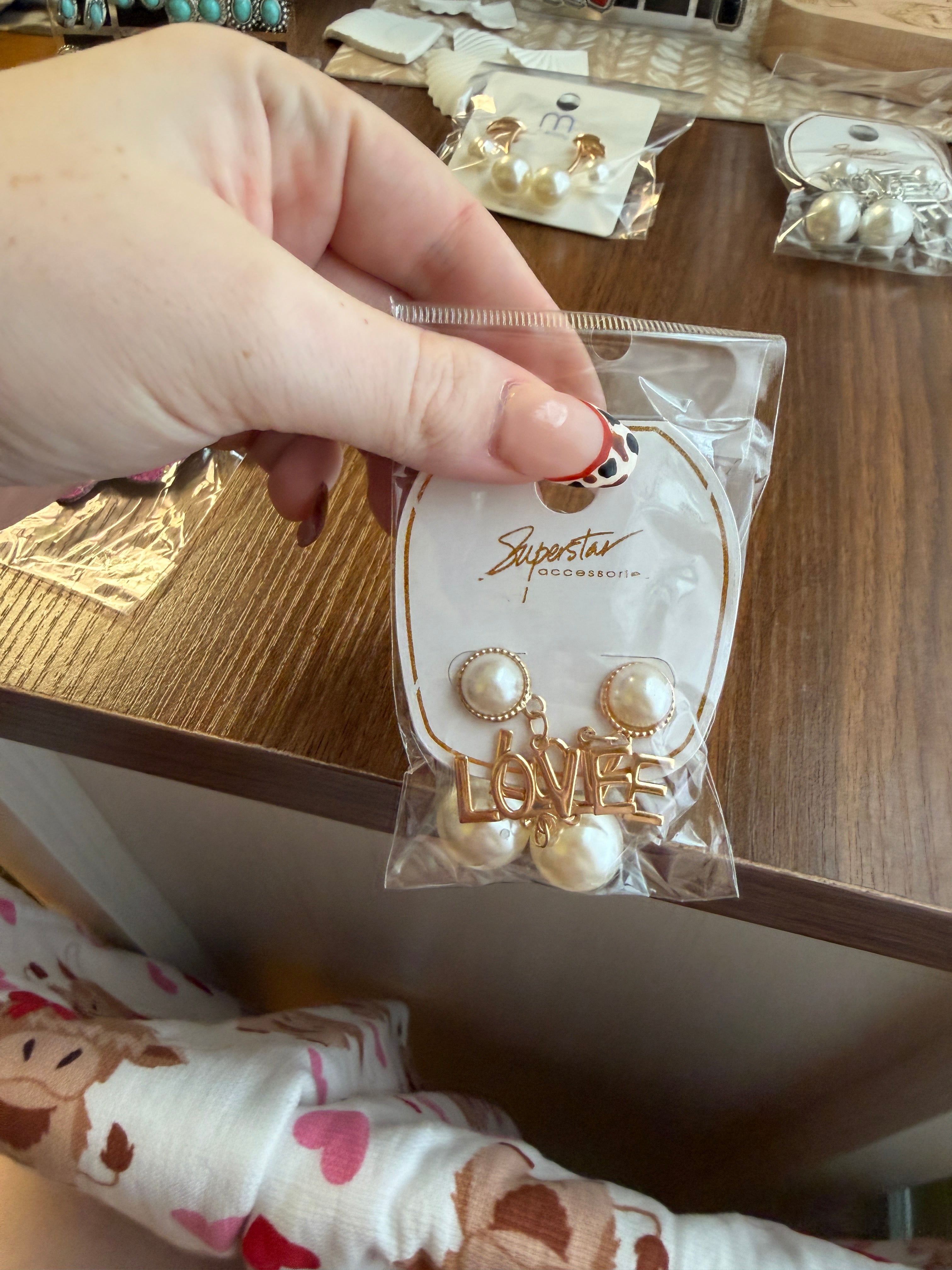 Hand holding a clear package with pearl earrings and a 'LOVE' tag on a wooden surface.