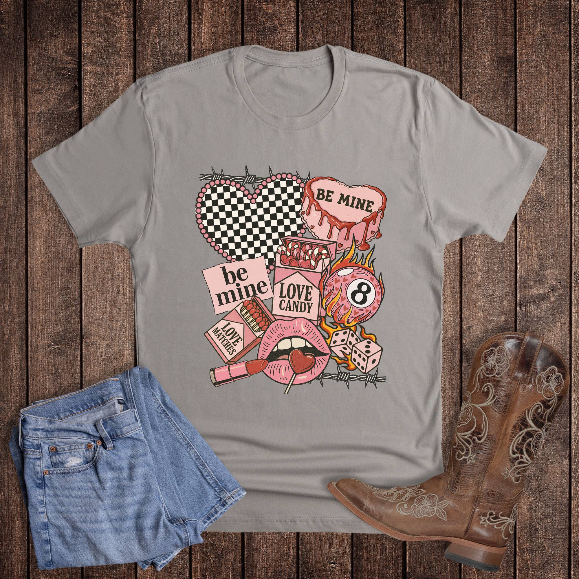 Gray t-shirt with colorful graphic design on a wooden surface with denim shorts and cowboy boots.