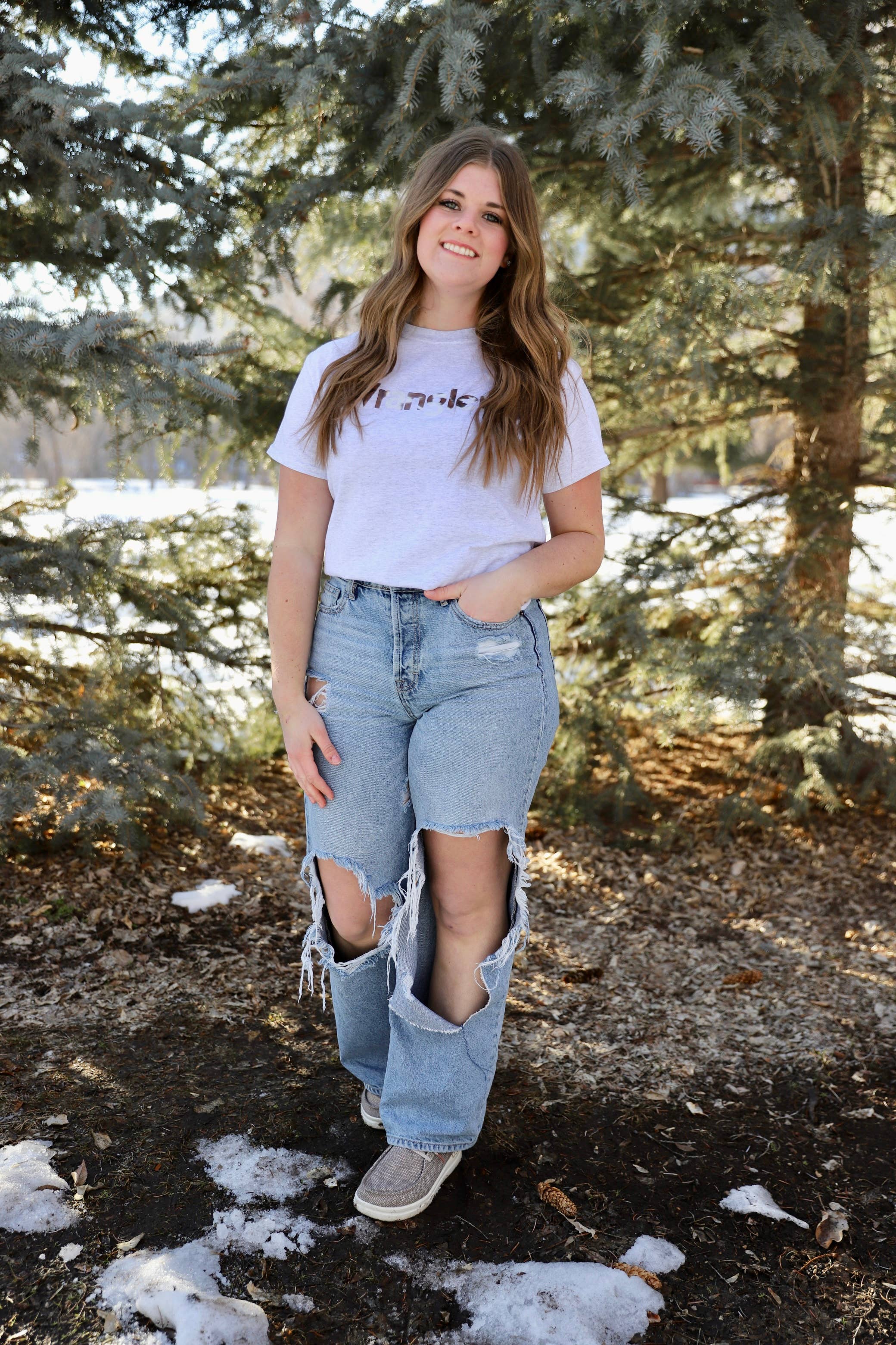 Person wearing a white t-shirt and ripped jeans standing in a snowy forest.