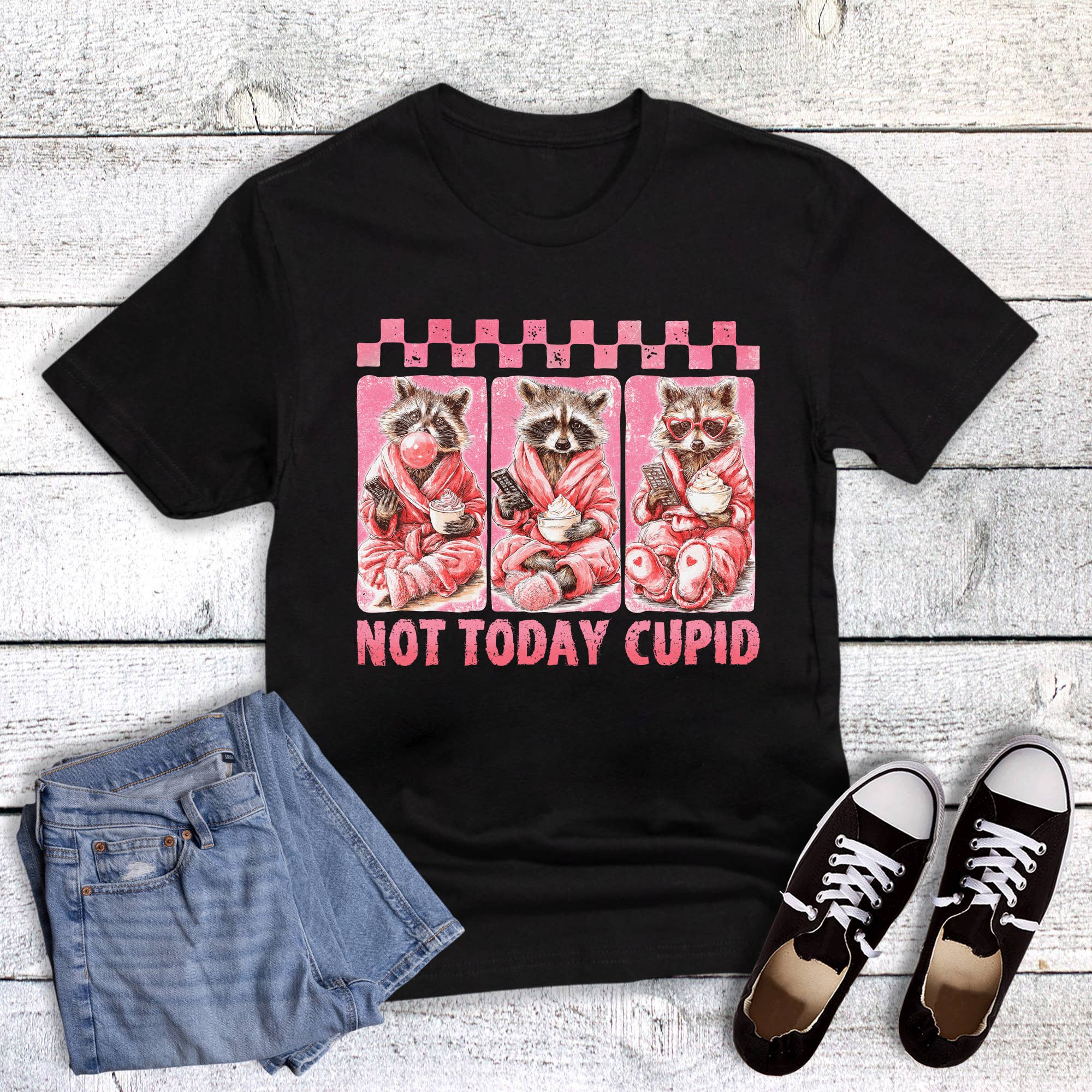 Black t-shirt with raccoon graphic and 'Not Today Cupid' text, paired with denim shorts and black sneakers on a wooden surface.